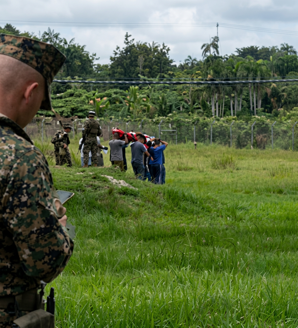 Un soldado observa desde el primer plano, mientras un grupo de personas en el fondo sostiene objetos sobre sus cabezas en un campo verde.